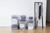 Several clear plastic storage boxes filled with folded clothes are stacked on a wooden floor against a white wall. A black garment rack with clothes hanging on it is to the right. Anteprima dell’immagine prodotto