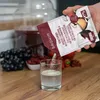 A person is pouring a wine yeast mixture from a packet into a glass of liquid, with grapes and other fruits in the background. Produktbild-Vorschau