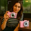 A woman is holding a pink digital camera in a cafe setting. Produktbild-Vorschau