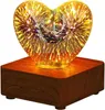 A heart-shaped glass lamp with a 3D firework effect sits on a wooden base. Produktbild-Vorschau