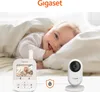 A baby is lying on a bed, with a baby monitor and camera shown in the foreground. Produktbild-Vorschau