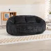 A dark gray bean bag sofa with two cushions is placed on a rug in a living room setting. A wooden cabinet with books and a camera is to the left, and a tall floor lamp is to the right.