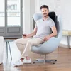 A man is sitting in a modern chair, holding a remote control, with a massage cushion on his neck and back. Produktbild-Vorschau
