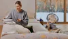A woman is sitting on a bed reading a book, while a baby is in a swing in the background near a window with a winter scene. Produktbild-Vorschau