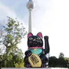 A black lucky cat figurine is placed in front of a tall tower and trees. Produktbild-Vorschau