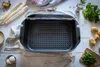 A top-down view of a rectangular roasting pan with a perforated insert and a glass lid, surrounded by ingredients like asparagus, potatoes, and herbs. Produktbild-Vorschau