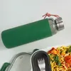A glass water bottle with a green silicone sleeve and a metal lid, placed next to a lunchbox with pasta. Anteprima dell’immagine prodotto