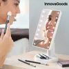 A woman applying makeup using a lighted mirror on a desk with makeup brushes and lipstick. Produktbild-Vorschau