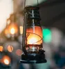 A close-up shot of a black kerosene lantern with a warm orange glow emanating from within, hanging in an outdoor setting with blurred lights in the background. Náhľad obrázku produktu