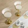 Two cream-colored bar stools with gold bases are shown on a light brown wooden floor, next to a white table with some decorative items. Voir les images du produit