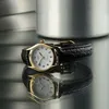 A Casio women's watch with a white face and gold-tone case, featuring a black leather strap, is shown on a reflective surface, with a blurred metallic background. Produktbild-Vorschau