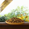 A wooden bowl filled with dried carob pieces sits on a wooden surface, with a blurred background of green foliage and yellow flowers. Produktbild-Vorschau