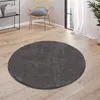 A round, dark gray rug is placed on a wooden floor in a room with a chair and a shelf in the background.
