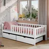 A white children's bed with pink bedding and a guardrail is shown in a room with a window and decorative wallpaper.