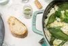 A close-up shot of a sage green Staub pot filled with risotto, garnished with fresh sage leaves and broccoli, next to a slice of bread and a small jar of herbs. Produktbild-Vorschau