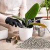 A person wearing black gloves is repotting a plant into a white pot, surrounded by gardening tools and a pile of substrate. Produktbild-Vorschau