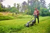 A man is mowing a lawn with a cordless lawnmower in a park-like setting. Produktbild-Vorschau