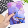 A hand holding a purple and blue marbled binder notebook with gold accents, placed on a desk with a laptop and a plant in the background. Voir les images du produit
