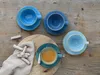 Four blue coffee cups and saucers are arranged on a wooden table, with some biscuits and a napkin on a wooden board. Náhľad obrázku produktu