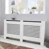 A white radiator cover with horizontal slats is shown in a room with a window and plants in the background. Podgląd zdjęcia produktu