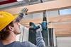A construction worker wearing a hard hat and safety glasses is using a cordless drill to attach a wooden beam to a ceiling structure. Náhled obrázku produktu