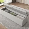 A light gray upholstered storage bench with its lid open, revealing stored items inside, placed on a rug in a room. Voir les images du produit