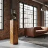 A modern living room with exposed brick walls, large windows, a leather sofa, and a wooden pedestal displaying a decorative item. Voir les images du produit