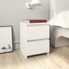 A white bedside table with two drawers is placed next to a bed. A red book rests on top of the table. A framed picture with two potted cacti is on the left. Produktbild-Vorschau