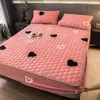 Rosa gesteppter Matratzenschoner mit schwarz-weißen Herzmustern, auf einem Bett mit passenden Kissenbezügen, eine Hand passt die Ecke an Náhled obrázku produktu