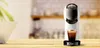 A white and black De'Longhi capsule coffee machine is shown on a wooden countertop with a cup of coffee in front of it. The background is blurred and appears to be a kitchen. Produktbild-Vorschau