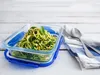 A glass food container filled with zucchini noodles and pesto, placed on a white wooden surface next to a folded napkin with cutlery. Anteprima dell’immagine prodotto