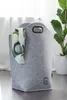 A grey felt laundry basket with a scarf draped over the side, placed on a wooden floor next to a potted plant. Produktbild-Vorschau