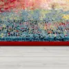 A close-up, low-angle view of the edge of a rug with a red border, placed on a light-colored wooden floor. Produktbild-Vorschau