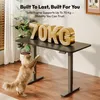 A desk with large gold letters spelling '70KG' on top, and a cat reaching up towards it. The desk is in a room with a plant and wooden flooring. Produktbild-Vorschau