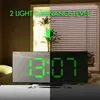 A digital alarm clock is shown on a table in front of a window with blinds. The clock displays the time and has a mirrored surface. Produktbild-Vorschau