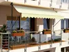 A balcony with a cream-colored awning extended, showing planters with flowers and a glass railing. Anteprima dell’immagine prodotto