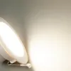 A close-up shot of a white LED downlight with a bright light emanating from it, casting a warm glow on a light gray wall. Produktbild-Vorschau