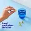 A hand holding a yellow capsule over a blue glass of water, with other capsules on a blue surface. Produktbild-Vorschau
