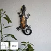 A solar-powered LED gecko decoration mounted on a textured wall, with plants in the foreground. Produktbild-Vorschau