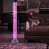 A tall, illuminated bubble lamp with pink water and decorative fish stands in a modern living room next to a brown leather sofa. Produktbild-Vorschau