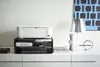 A white printer sits on top of a black multi-tiered shelf unit, which holds paper and ink cartridges. A laptop is on the bottom shelf. A lamp and decorative vase are on the right side of the shelf. Produktbild-Vorschau