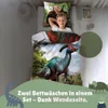 A young boy is lying on a bed with dinosaur-themed bedding. The bedding features detailed illustrations of dinosaurs and a natural landscape. Produktbild-Vorschau