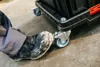 A person's foot in a dirty work boot is pressing on the wheel of a black tool case on a concrete floor. Náhled obrázku produktu