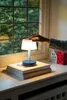 A hand touches a small table lamp on a windowsill, with decorative houses and a window view in the background. Produktbild-Vorschau
