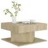 A modern, square coffee table with a unique layered design, presented against a clean white background. The table has a light wood grain finish and is styled with a vase of green leaves, a magazine, and two white coffee cups. Voir les images du produit
