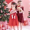 Two young girls in red dresses holding baby dolls in front of a decorated Christmas tree. Produktbild-Vorschau
