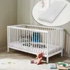 A white baby crib with a mattress and pillows inside, placed on a wooden floor with a rug and toys nearby. A close-up of a mattress is shown in an inset circle at the top. Produktbild-Vorschau