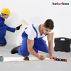 Two construction workers are working on a white wall. One is painting with a roller, and the other is kneeling and measuring something on the floor with a tape measure and a level. Náhľad obrázku produktu