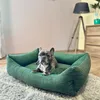 A French bulldog is resting in a green dog bed on a fluffy gray rug. In the background, there is a pink wall, a wooden dresser, and a floor lamp. Náhled obrázku produktu