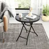 A round black folding table with a glass top is placed on a rug next to a chair, with a plant and window in the background. Produktbild-Vorschau